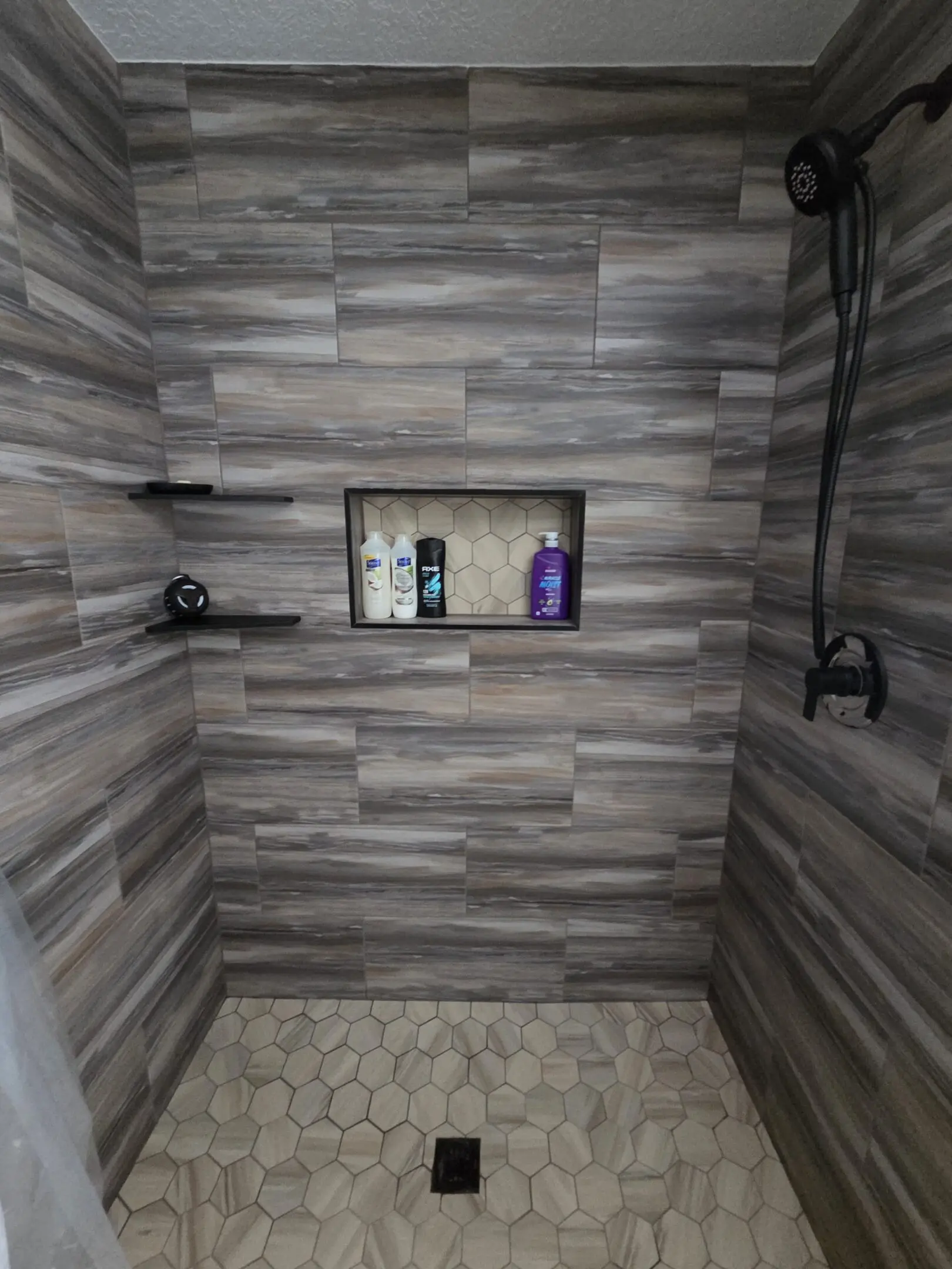 Modern shower with gray tile walls and built-in shelf holding bottles.